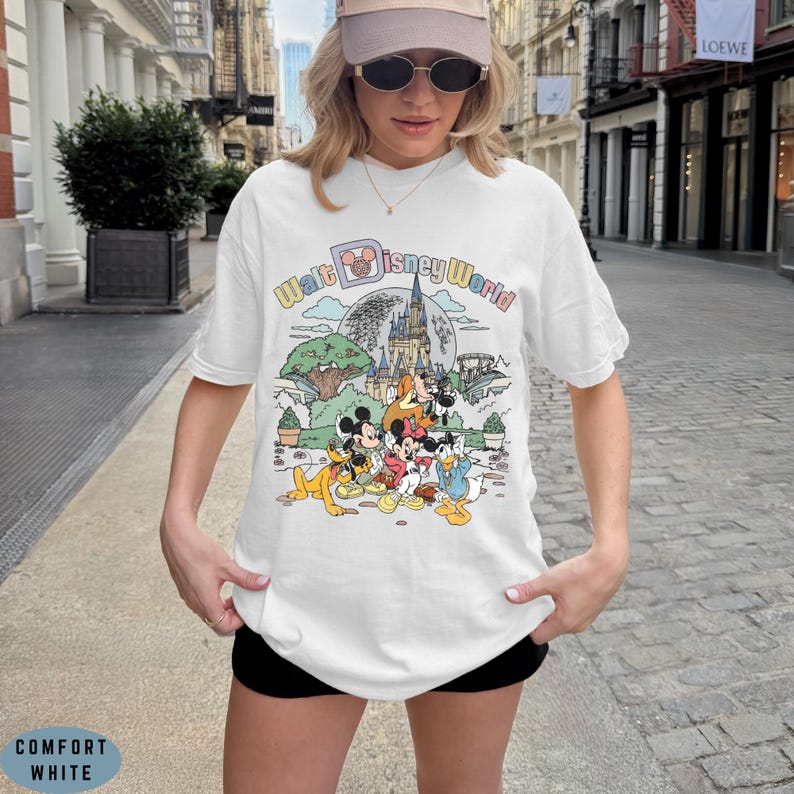 May include: White t-shirt with a colorful graphic featuring Disney characters and the text "Walt Disney World." The design includes Mickey Mouse, Minnie Mouse, Donald Duck, and other characters in front of a castle and a tree.