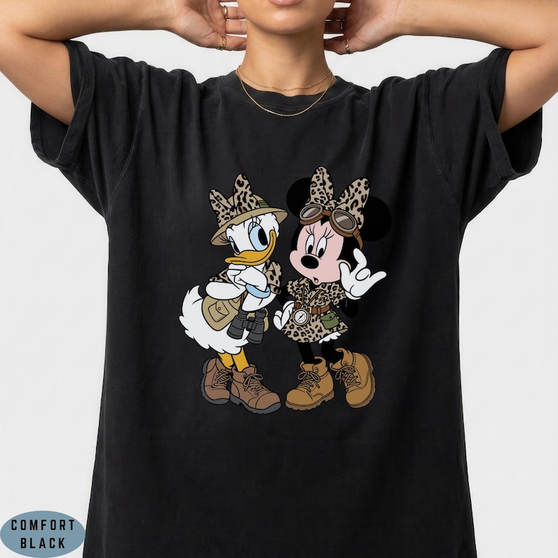 K&ouml;nnte beinhalten: Ein schwarzes T-Shirt mit einem Grafikdruck von Daisy Duck und Minnie Mouse in Safari-Kleidung, bestehend aus H&uuml;ten mit Leopardenmuster, Westen und braunen Stiefeln. Die Figuren sind im Cartoon-Stil nebeneinander stehend abgebildet.