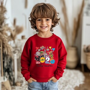 May include: A red sweatshirt featuring a colorful graphic with the text "2020 Family Vacation" and various Disney-themed icons. The design includes Mickey and Minnie Mouse, a castle, and other park elements. The sweatshirt is worn by a child.