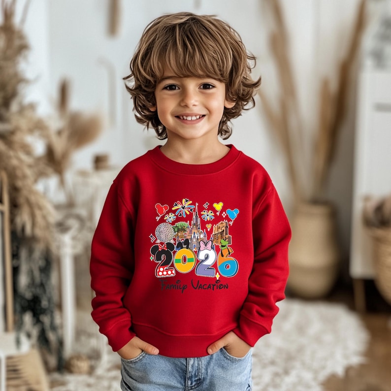 May include: A red sweatshirt featuring a colorful graphic with the text "2020 Family Vacation" and various Disney-themed icons. The design includes Mickey and Minnie Mouse, a castle, and other park elements. The sweatshirt is worn by a child.