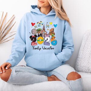 May include: Light blue hoodie with a colorful graphic featuring the numbers "2026" and the words "Family Vacation." The design includes cartoon images of a castle, a golf ball, and other theme park elements. The hoodie has a front pocket and a hood.