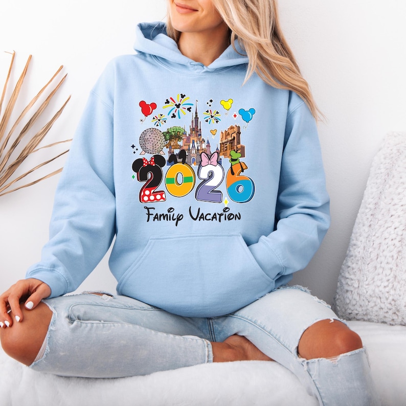 May include: Light blue hoodie with a colorful graphic featuring the numbers "2026" and the words "Family Vacation." The design includes cartoon images of a castle, a golf ball, and other theme park elements. The hoodie has a front pocket and a hood.