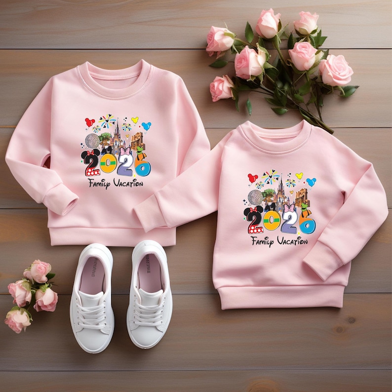 May include: Two pink sweatshirts with a colorful graphic featuring Mickey and Minnie Mouse, the number 2020, and the words "Family Vacation." White sneakers are at the bottom. A bouquet of pink roses rests on a wooden surface.