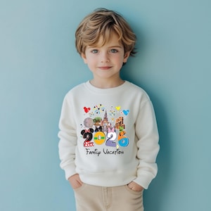 May include: A cream-colored sweatshirt featuring a colorful graphic with the text "2020 Family Vacation." The graphic includes images of a castle, a mouse, and other cartoon characters. The sweatshirt is worn by a young child.