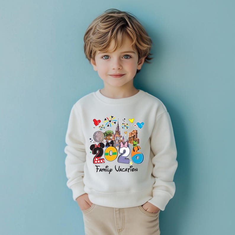 May include: A cream-colored sweatshirt featuring a colorful graphic with the text "2020 Family Vacation." The graphic includes images of a castle, a mouse, and other cartoon characters. The sweatshirt is worn by a young child.