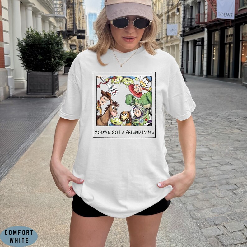 May include: A white short-sleeve t-shirt featuring a graphic print of a square polaroid-style frame containing various animated toy characters. Below the illustration, the text reads YOU'VE GOT A FRIEND IN ME in a simple black sans-serif font.