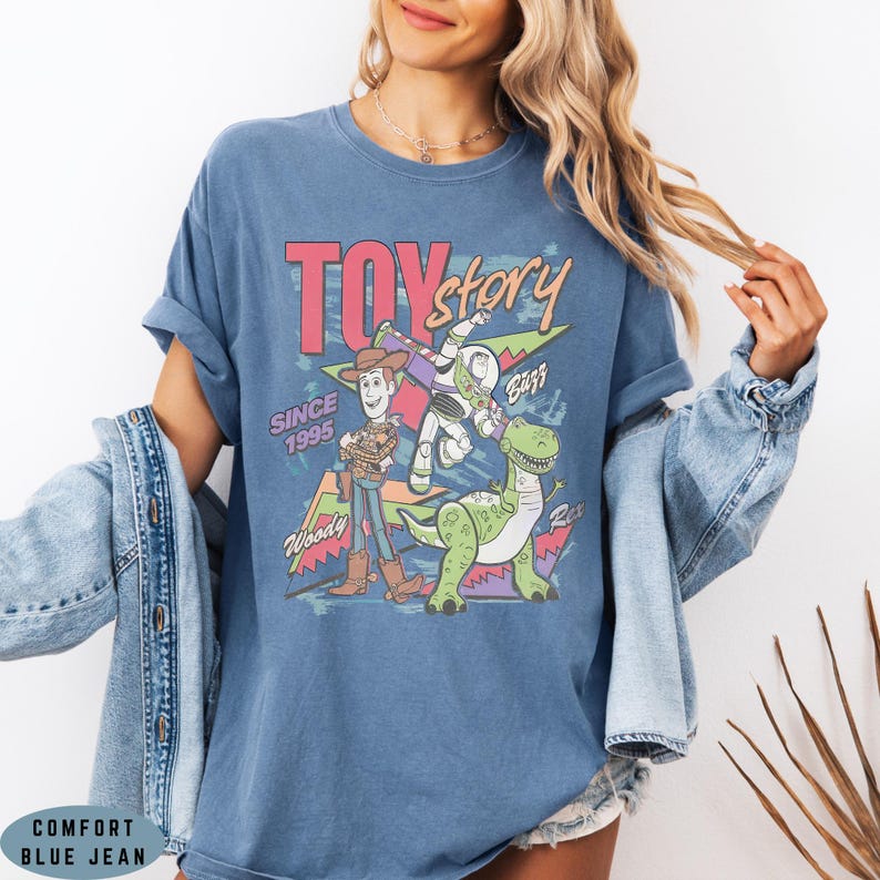 May include: A blue short-sleeve t-shirt featuring a graphic print of Woody, Buzz Lightyear, and Rex from Toy Story. The design includes the text "TOY story", "SINCE 1995", "Woody", "Buzz", and "Rex" with colourful geometric shapes in the background.