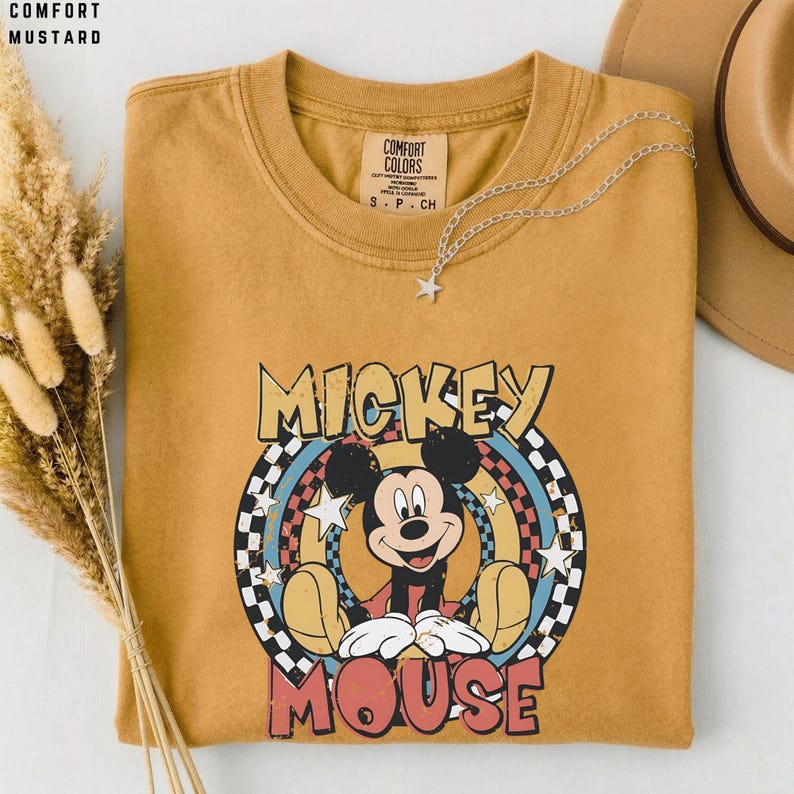 Puede incluir: Una camiseta de cuello redondo color amarillo mostaza con un gr&aacute;fico de Mickey Mouse centrado en un dise&ntilde;o circular. El gr&aacute;fico incluye un borde a cuadros, estrellas y el texto MICKEY MOUSE. La camiseta tiene una etiqueta de Comfort Colors.