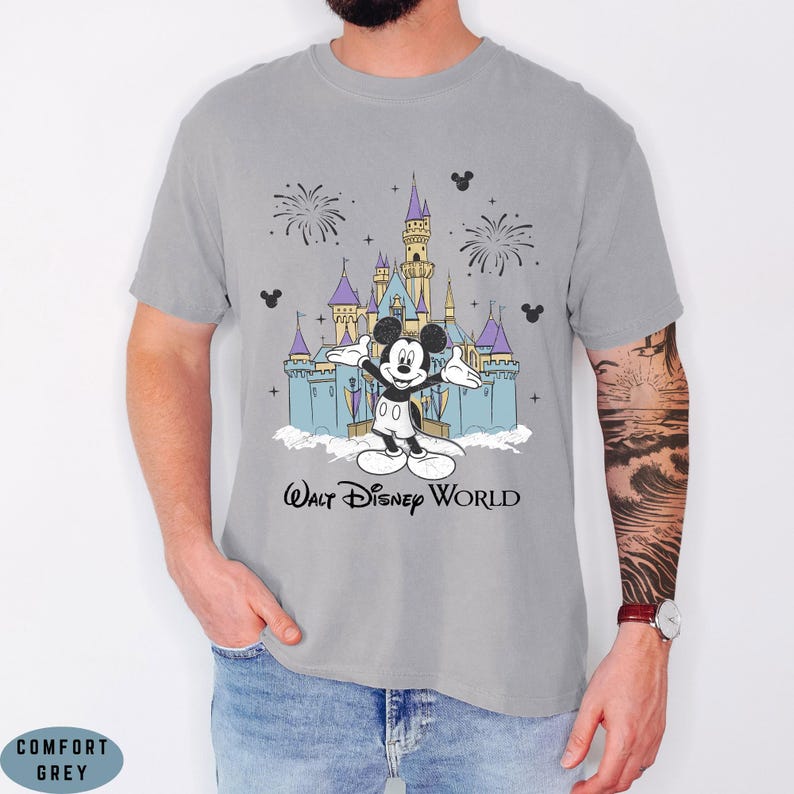 Op de afbeelding: Een grijs T-shirt met een afbeelding van Mickey Mouse voor het kasteel van Assepoester in Walt Disney World. Het ontwerp bevat vuurwerk en de tekst "Walt Disney World". Het shirt is gelabeld "Comfort Grey".