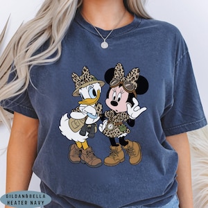 K&ouml;nnte beinhalten: Ein kurz&auml;rmeliges T-Shirt in Marineblau mit einem Grafikdruck von zwei Zeichentrickfiguren in Safari-Ausr&uuml;stung. Beide Figuren tragen H&uuml;te und Kleidung mit Leopardenmuster, braune Stiefel sowie Accessoires wie Ferngl&auml;ser, Brillen und Kompass.