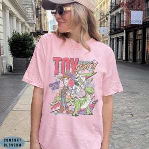 May include: A light pink short-sleeve t-shirt featuring a graphic print of Toy Story characters Woody, Buzz Lightyear, and Rex. The design includes the text Toy Story, Since 1995, Woody, Buzz, and Rex with colourful geometric shapes in the background.