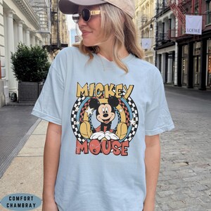 Puede incluir: Una camiseta de manga corta de color azul claro con un gr&aacute;fico de Mickey Mouse centrado en un marco circular. El dise&ntilde;o incluye un borde a cuadros blanco y negro, detalles en amarillo y rojo, estrellas blancas y el texto MICKEY MOUSE en negrita.