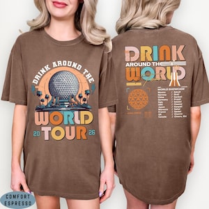 May include: Brown t-shirt with a graphic design featuring the text "DRINK AROUND THE WORLD TOUR 26". The front design includes a sphere and the back design lists countries and phrases. The shirt is labeled "COMFORT ESPRESSO".