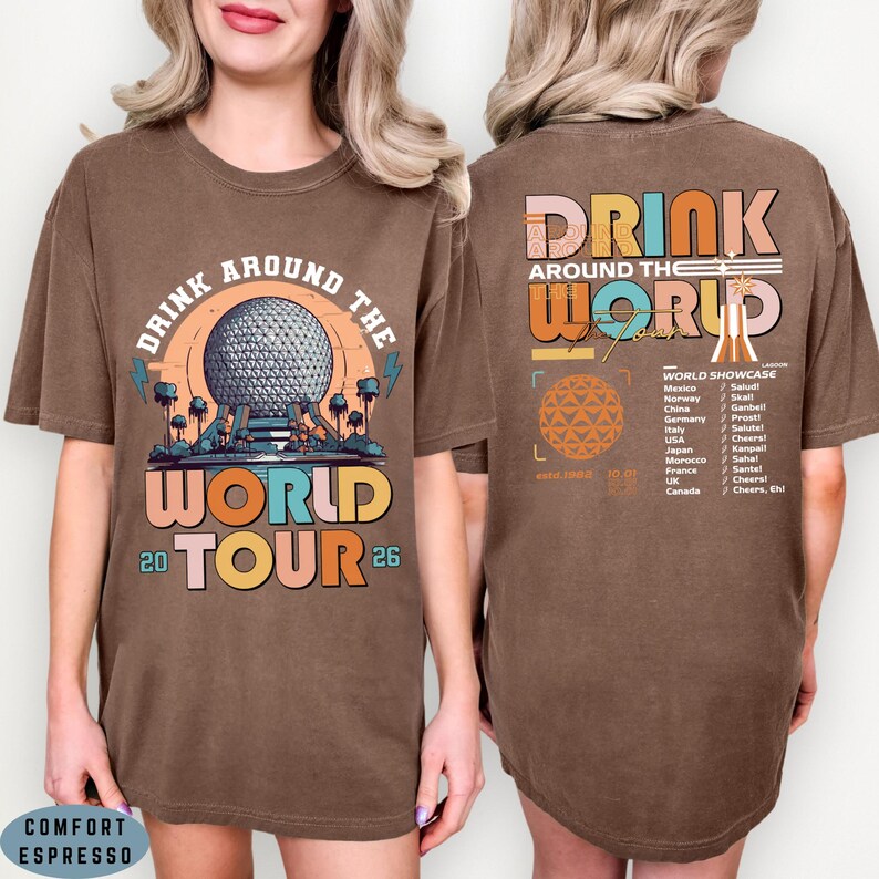 May include: Brown t-shirt with a graphic design featuring the text "DRINK AROUND THE WORLD TOUR 26". The front design includes a sphere and the back design lists countries and phrases. The shirt is labeled "COMFORT ESPRESSO".