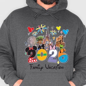 May include: Dark gray hoodie with a colorful graphic design. The design features the numbers "2026" with Mickey and Minnie Mouse ears, a castle, and the words "Family Vacation". Balloons and fireworks are also included in the design.