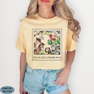 May include: A light yellow short-sleeve t-shirt featuring a square graphic print of various animated characters from Toy Story. Below the characters, the text reads YOU'VE GOT A FRIEND IN ME in black capital letters within the square frame.