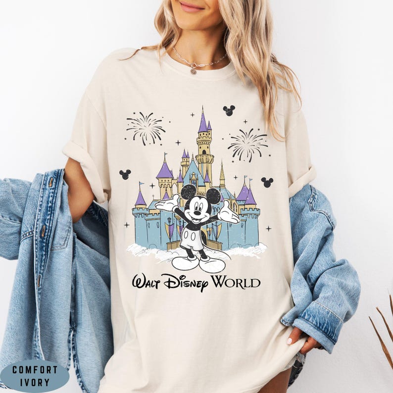 Op de afbeelding: Ivoor kleurig T-shirt met een afbeelding van Mickey Mouse voor het kasteel van Assepoester in Walt Disney World. Het ontwerp bevat vuurwerk en de tekst "Walt Disney World". Het shirt is gelabeld "Comfort Ivory".