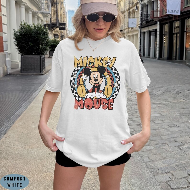 Puede incluir: Una camiseta blanca de manga corta con un estampado gr&aacute;fico de Mickey Mouse centrado en un marco circular. El dise&ntilde;o incluye un borde a cuadros blanco y negro, detalles en amarillo y el texto Mickey Mouse en una fuente roja desgastada.