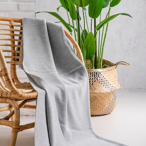 May include: A light gray knitted throw blanket draped over a wicker chair. A large potted plant with green leaves sits in a woven basket in the background. The blanket has a textured surface and a finished edge.