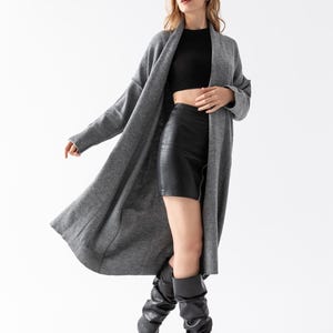 May include: A long, gray cardigan sweater with a flowing design. The model is wearing a black crop top, black leather mini skirt, and black knee-high heeled boots. The outfit is set against a white background.