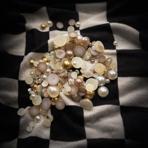 May include: A collection of assorted beads and embellishments in various sizes and colors, including gold, cream, and pearl, are scattered on a black and white checkered fabric. The beads appear to be made of different materials, creating a textured and visually appealing display.