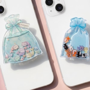 May include: Two white smartphones each with a decorative phone grip. One grip features a blue bag design with cartoon fish and the text "PURRMAIDS". The other grip has a similar design with different fish.