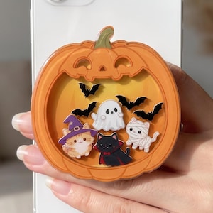 Può includere: Un supporto circolare per telefono a forma di zucca arancione con interno giallo. All'interno ci sono piccole figure di un gatto con un cappello da strega viola, un fantasma bianco, un gatto nero con mantello da vampiro, un gatto mummia e pipistrelli.
