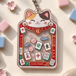 May include: A clear acrylic keychain featuring a calico cat head at the top and a rectangular frame containing floating mahjong tiles, fish, and paw print shapes. The frame is decorated with red, gold, and green patterns and the text Lucky Charm.