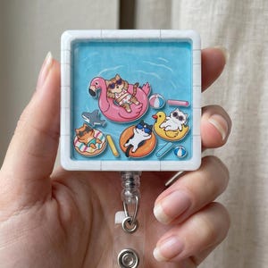 Puede incluir: Un carrete retráctil de insignia cuadrado blanco con una escena de piscina con gatos de dibujos animados en flotadores. Los gatos llevan gafas de sol y se relajan en un flamenco, un pato y otros flotadores. El carrete tiene una correa de plástico transparente y un clip de metal.