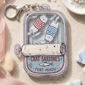 May include: A novelty keychain featuring a sardine can design with two cartoon cats wearing striped shirts and several fish. The can reads "CHAT SARDINES - PORT MIAOU" in blue and red. The keychain has a silver clasp.