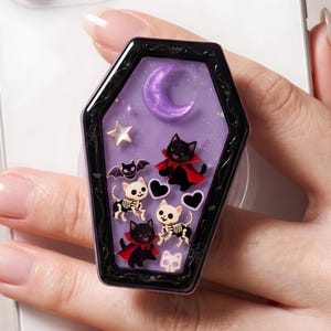 May include: A coffin-shaped phone grip featuring a purple background with a crescent moon, a star, and black heart shapes. The design includes two black cats wearing red capes, two skeleton cats, and a small bat, all encased in a black frame.