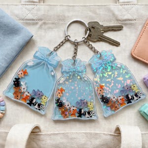 Peut inclure: Trois porte-clés avec un design de sac bleu rempli de petits poissons colorés. Chaque porte-clés a un nœud bleu clair en haut et une chaîne argentée. L'arrière-plan comprend des clés, un portefeuille et un bracelet.