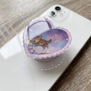 May include: A heart-shaped phone grip with a scalloped edge featuring a glossy finish. The design displays a brown tabby cat floating in a purple and blue galaxy background with a crescent moon, a planet with rings, and several small stars.