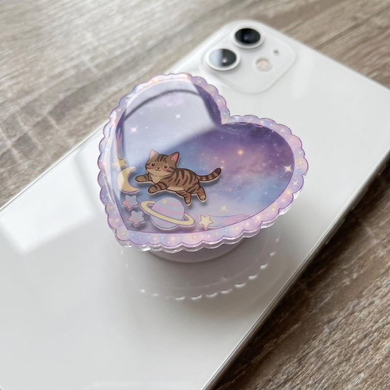 May include: A heart-shaped phone grip with a scalloped edge featuring a glossy finish. The design displays a brown tabby cat floating in a purple and blue galaxy background with a crescent moon, a planet with rings, and several small stars.