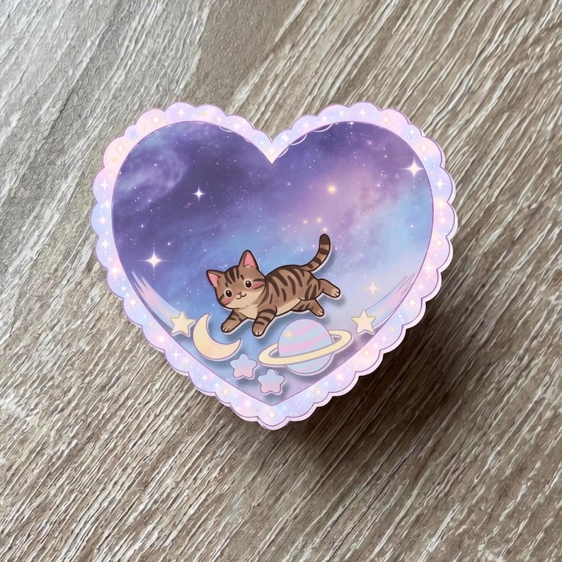 May include: A heart-shaped sticker with a scalloped edge featuring a purple and blue galaxy background. A brown tabby cat is illustrated in the center, floating above a small crescent moon, a ringed planet, and several small stars.