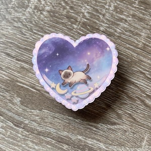 May include: A heart-shaped sticker with a scalloped edge featuring a Siamese cat floating in a purple and blue galaxy background. The design includes a small yellow crescent moon, a pastel ringed planet, and several small stars scattered throughout.