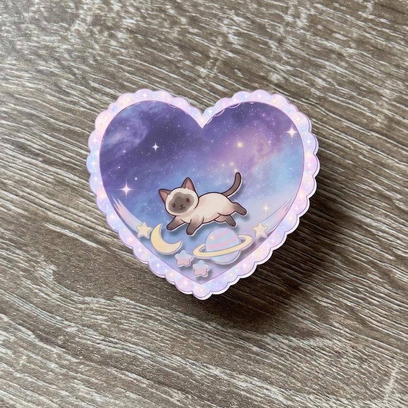 May include: A heart-shaped sticker with a scalloped edge featuring a Siamese cat floating in a purple and blue galaxy background. The design includes a small yellow crescent moon, a pastel ringed planet, and several small stars scattered throughout.