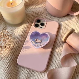 May include: A light pink smartphone case featuring a heart-shaped grip attachment. The grip displays a graphic of a Siamese cat floating in a purple and blue starry space scene with a crescent moon, a planet with rings, and small white stars.