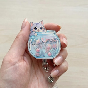 May include: A cute cat badge reel with a blue and white striped cat peeking over the edge of a fishbowl. The fishbowl contains jellyfish, coral, and seashells. The reel has a retractable cord and a metal clip.