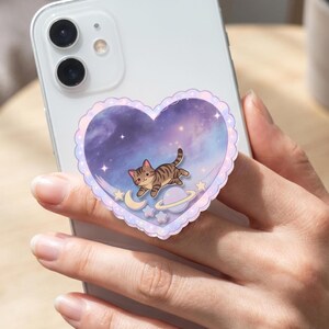 May include: A heart-shaped phone grip attached to the back of a white smartphone. The grip features a scalloped edge and a purple galaxy background with a brown tabby cat, a small crescent moon, a planet with rings, and several stars.