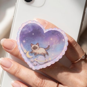 May include: A heart-shaped phone grip featuring a scalloped edge and a purple and blue galaxy background. The design includes a small Siamese cat floating in space, accompanied by a yellow crescent moon, a ringed planet, and several small stars.