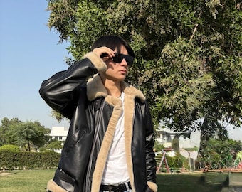 May include: Black leather jacket with a beige shearling lining and trim. The jacket features a wide collar, a belt at the waist, and a classic aviator style. The person is wearing sunglasses and light blue jeans.