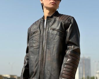 May include: A dark brown leather jacket with a zip-up front, two chest pockets, and ribbed detailing on the shoulders and sleeves. The jacket is worn with blue jeans and sunglasses.