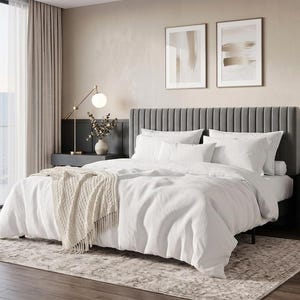 May include: A bedroom set featuring a grey upholstered headboard with vertical channel tufting, a white duvet cover, white pillows, and a cream-colored knit throw blanket with fringe, arranged on a bed frame with a neutral-toned area rug.
