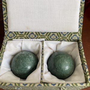 Può includere: Due pietre sferiche verdi e lisce sono all'interno di una scatola divisa. La scatola ha un esterno verde e blu a fantasia e un interno bianco. Le pietre sono immerse in tessuto bianco.