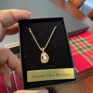 Puede incluir: Un collar dorado con un colgante de ópalo genuino, presentado en una caja negra. El colgante presenta una piedra de ópalo nacarada engastada en un marco dorado texturizado. La caja incluye una etiqueta dorada que dice "Genuine Opal Necklace".