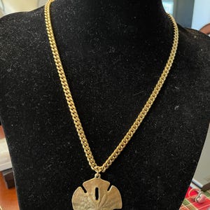 Puede incluir: Un collar dorado con un gran colgante de dólar de arena. La cadena es de estilo eslabón grueso. El colgante tiene un diseño detallado y se exhibe sobre un busto de terciopelo negro.