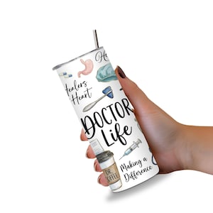 May include: A white tumbler with a silver straw, held in a hand. The tumbler is decorated with medical-themed illustrations, including a stomach, pills, and a reflex hammer. The words "DOCTOR LIFE" are prominently displayed, along with "Healers Heart" and "Making a Difference."