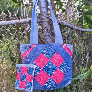 May include: A blue denim tote bag featuring a quilted patchwork design of red and blue paisley fabric squares. The bag includes two shoulder straps and a matching small zippered pouch attached by a silver metal clip and a short denim wrist strap.