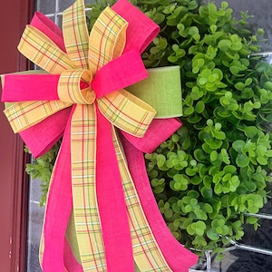 May include: A decorative green wreath with a large bow. The bow is made of bright pink, yellow plaid, and green ribbons. The wreath is composed of small, round green leaves, creating a colourful and cheerful decorative piece for the home.